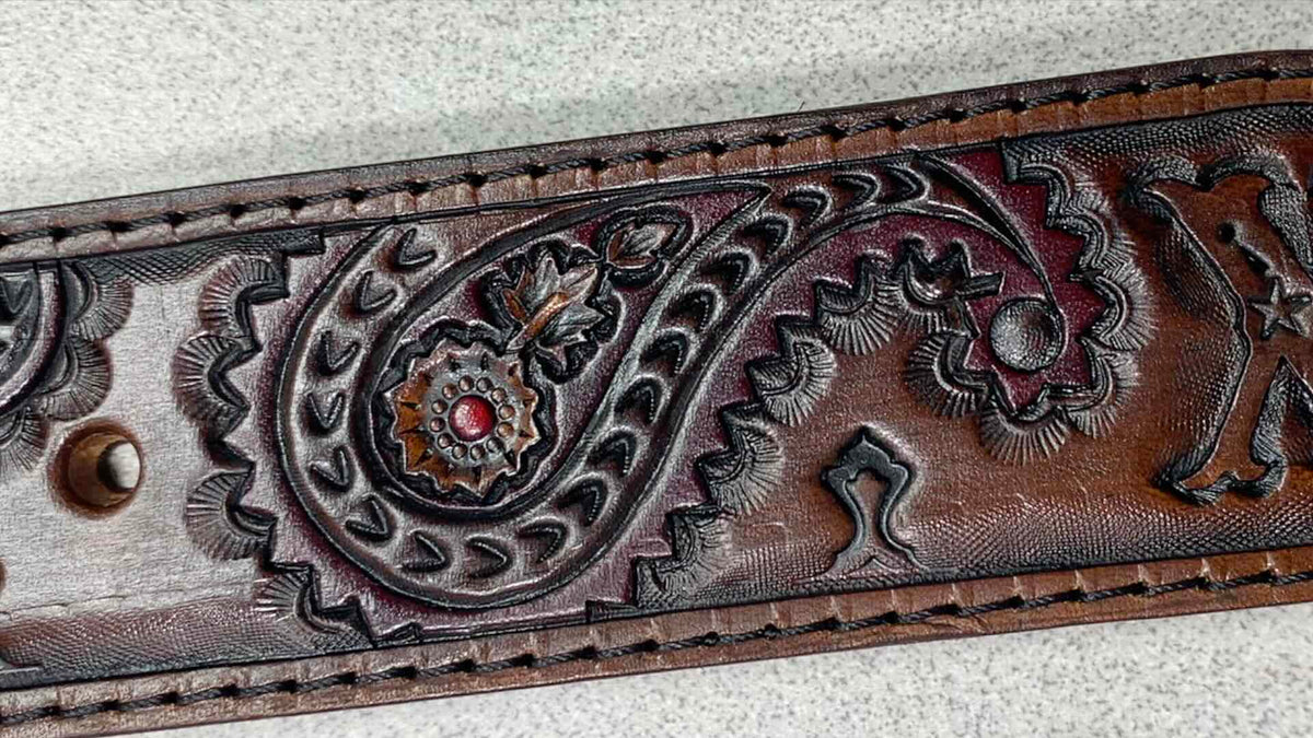 A close-up of a brown tooled leather belt featuring an embossed paisley pattern with a small red gemstone accent in the center, resting on a light gray textured surface.